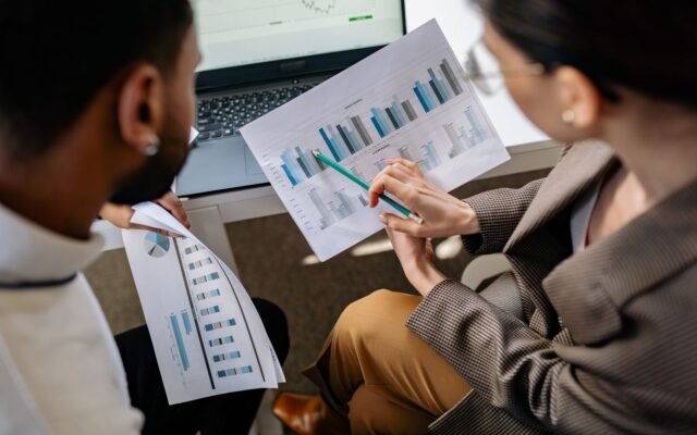 Two colleagues going through printed bar charts together at a desk, with a laptop open behind them